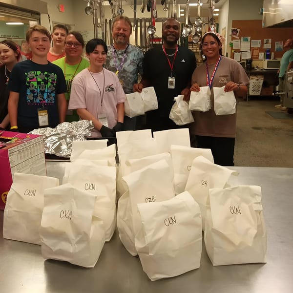 A group of smiling cafe staff and volunteers stand behind a table filled with white to-go bags.