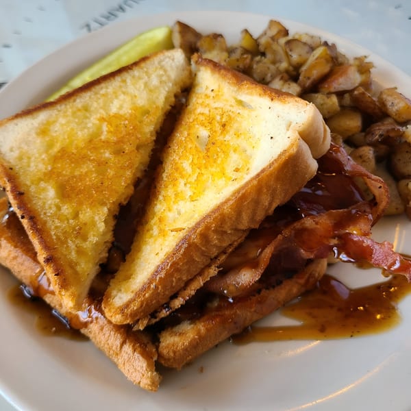 Bacon and toast sandwich with syrup, a pickle spear, and home fries on a white plate.