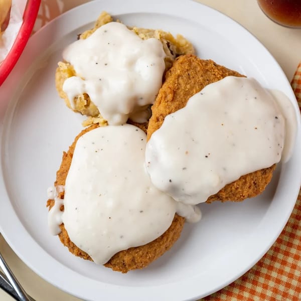 Three pieces of golden-fried chicken or steak, generously covered in creamy white gravy, on a white plate.
