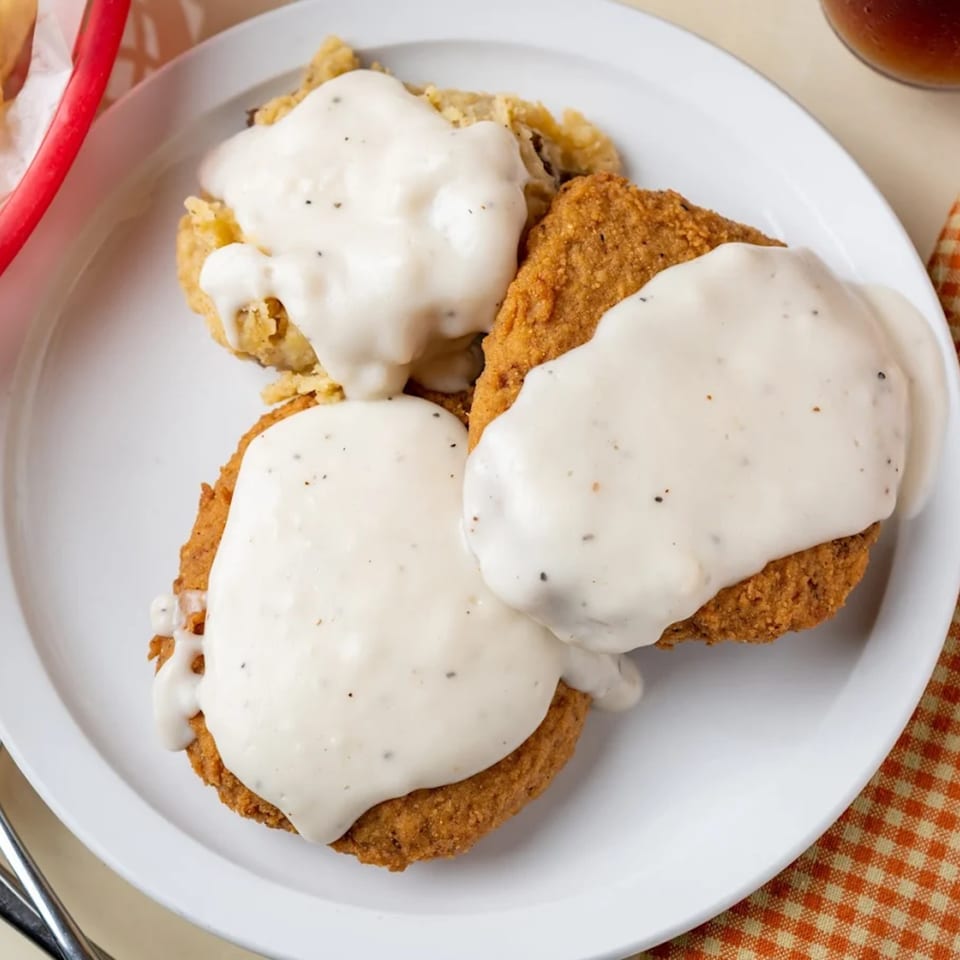 Three pieces of golden-fried chicken or steak, generously covered in creamy white gravy, on a white plate.