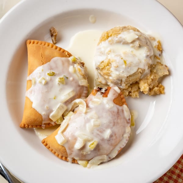 Two golden-brown savory pastries covered in creamy white gravy with a side of stuffing on a white plate.