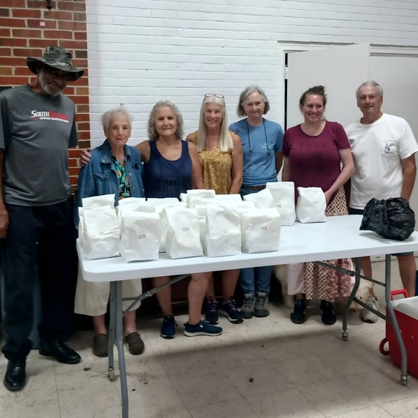 Seven people stand behind a table laden with white paper bags, likely meals from Time To Eat Cafe.