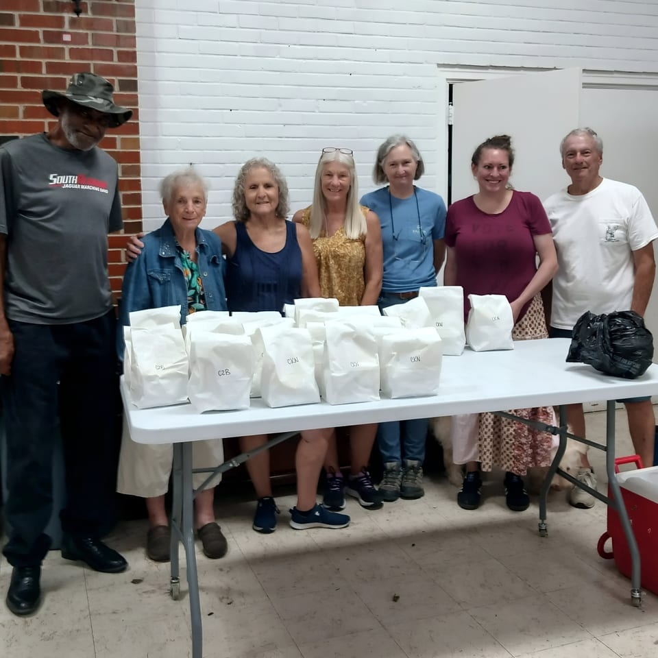 Seven people stand behind a table laden with white paper bags, likely meals from Time To Eat Cafe.