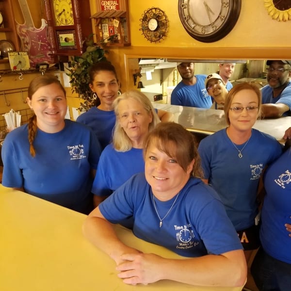 Smiling Time To Eat Cafe staff in blue shirts pose behind and over a counter.