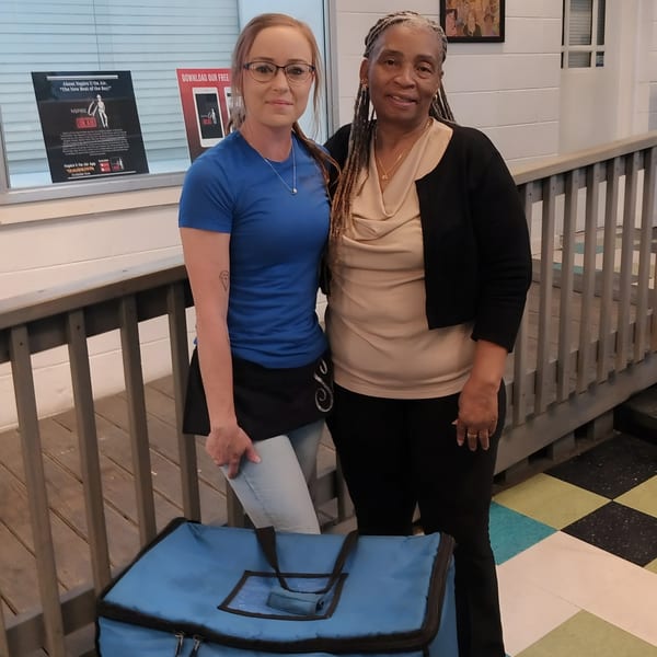 Two women, one in a blue shirt and the other in a black cardigan, stand next to a blue delivery bag.
