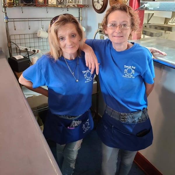 Two smiling women in blue Time To Eat Cafe shirts and aprons stand together behind a counter.